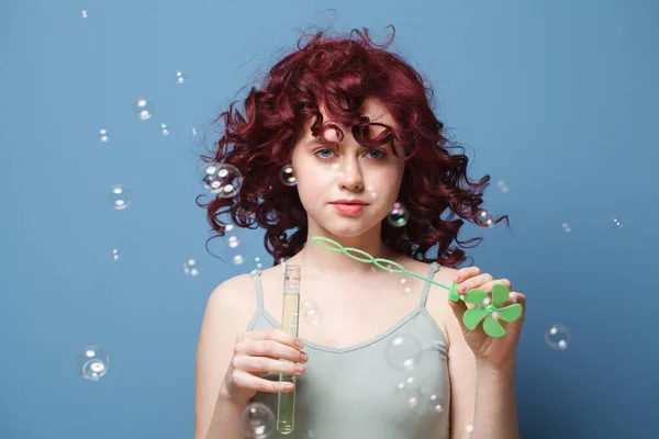 Beautiful woman with red curly hair blowing soap bubbles on blue background