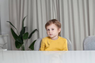 Happy child boy with brown hair in yellow sweater sitting by the table on beige curtain background
