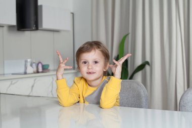 Portrait of cute child boy in yellow sweater sitting and raising his hands up on white kitchen background