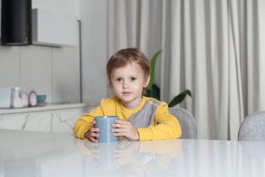Happy cute child boy sitting by the table and holding grey cup on white curtain background