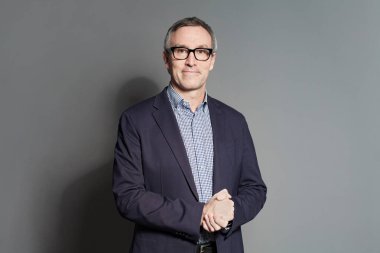 Takım elbiseli, mavi beyaz gömlekli ve gözlüklü, kendine güvenen bir iş adamı gri stüdyo arka planında duruyor.