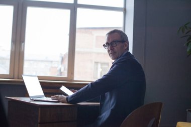 Masanın yanında oturan, dizüstü bilgisayarda çalışan ve cep telefonunu pencereye dayayan gri gözlükle yaşlı başarılı bir işadamı.