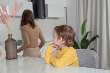 Kahverengi saçlı, sarı kazaklı, stüdyo mutfağında oturan ve annesine bakan mutlu çocuk.