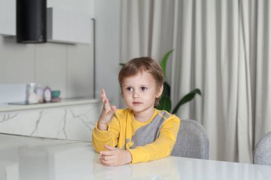Sarı süveterli şirin çocuğun portresi masada oturuyor ve elini beyaz stüdyo arka planında kaldırıyor.