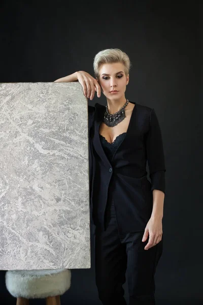 穿着黑色西服的漂亮女设计师站在黑色背景的灰色画架旁边 — 图库照片