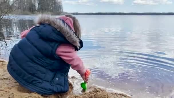 湖のそばで遊ぶ少女は緑のシャベルで砂を取ります — ストック動画