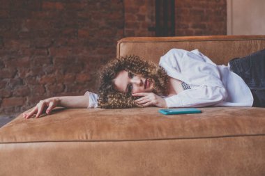 Beautiful glamorous woman in white shirt with curly hair resting on the sofa with green mobile phone near