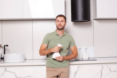 Casual attractive man cleaning kitchen and holding sptay bottle with blank label for advertising