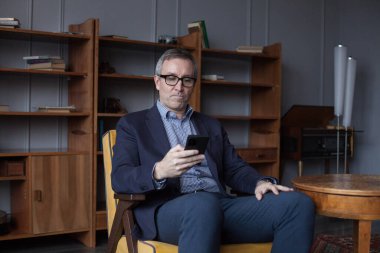 Confident elderly businessman in blue suit and glasses working and looking on his mobile phone indoor on office wall with boockshelf 