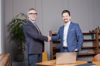 Takım elbiseli iki mutlu iş adamı ofiste tokalaşıyor, iş başarıya ulaşıyor.