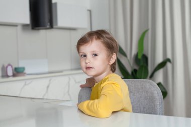 Kahverengi saçlı, sarı kazaklı mutlu çocuk stüdyo mutfağında yan oturuyor.