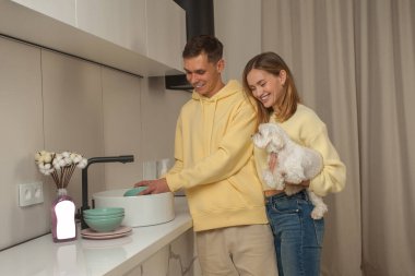 Bulaşıkları lavaboda yıkayan adam ve elinde küçük beyaz bir köpek tutan kadın, masadaki boş etiketle bulaşık deterjanı.