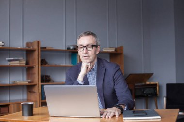 Gözlüklü ve takım elbiseli yetişkin işadamının portresi ofisinde dizüstü bilgisayarda oturuyor ve çalışıyor.