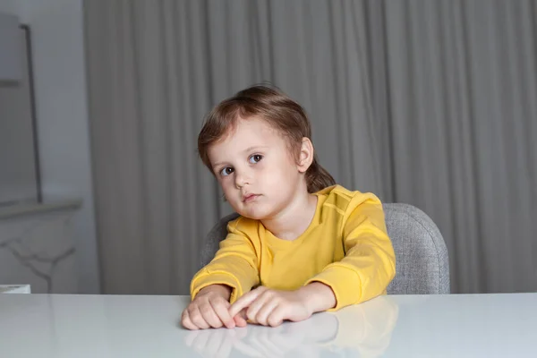 一个有着棕色头发 穿着黄色毛衣的多愁善感的小男孩坐在桌旁 看着摄像机 拉着双手 — 图库照片