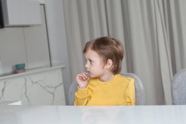 Kahverengi saçlı, sarı kazaklı dalgın çocuk bej perde arkasında masanın yanında oturuyor.