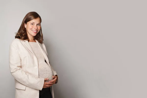 美しいです妊娠中の女性抱擁彼女のおなか 笑顔と立っていますとともに灰色のスタジオの壁の背景 — ストック写真