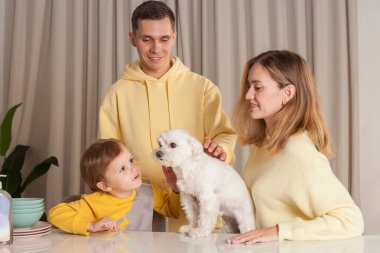 Sarı süveterli, çocuklu, beyaz köpekli sevimli ebeveynler masanın üzerinde üç soyut plastik şişe.