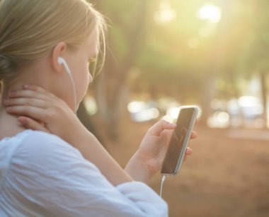 Beyaz gömlekli genç bir kızın portresi kulaklıkla müzik dinliyor ve arka planda ve güneş ışığında cep telefonuyla yazı yazıyor.