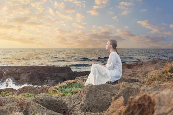 Genç kız mavi denizin yanındaki kayalarda oturuyor.