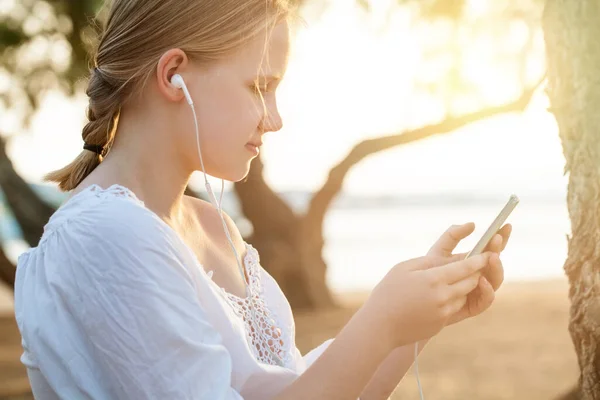 stock image Smiling blonde teenage girl in headphones holding mobile phone and standing sideways on the background of sea