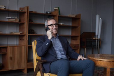 Yaşlı mutlu iş adamının portresi, gri saçlı, takım elbiseli, gözlüklü, gülümseyen ve gri arka planda cep telefonuyla konuşan gri kitaplıklı.