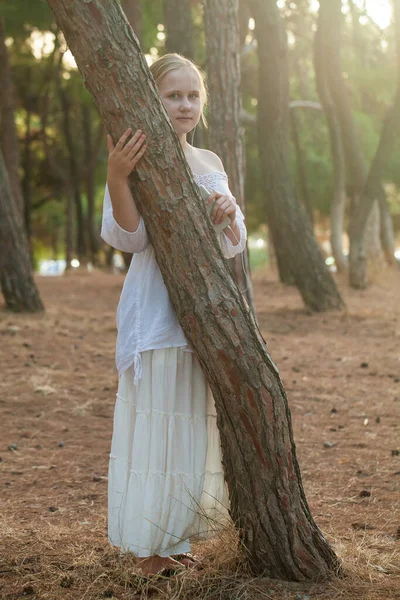 穿着白衬衫的金发少女站在树旁的森林中 — 图库照片