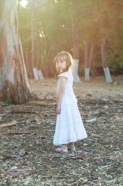 Beyaz elbiseli esmer bir kız çocuğu güneşli bir parkın arka planında dikiliyor.