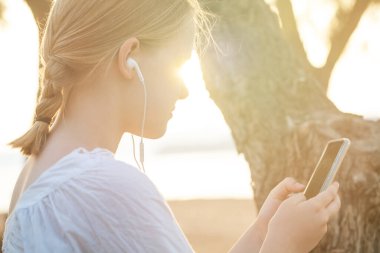 Kulaklıklı mutlu, gülümseyen genç bir kızın portresi. Elinde cep telefonu, arka planda dikiliyor.