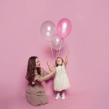 Pembe stüdyo geçmişinde pembe balonu olan neşeli, sevimli bir anne ve kız. Genç anne ve tatlı kız bebek portresi.