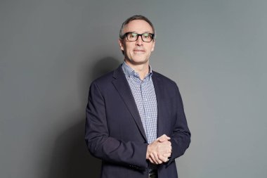 Takım elbiseli, kareli gömlekli ve gözlüklü yetişkin işadamının portresi gri stüdyo arka planında kameraya gururla bakıyor.