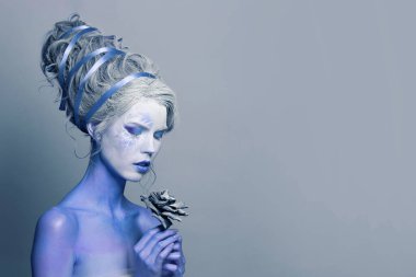 Beyaz yüzlü, sahne makyajlı ve vücutta mavi derili gizemli mükemmel kadın model gri arka planda siyah gül çiçeği tutuyor.