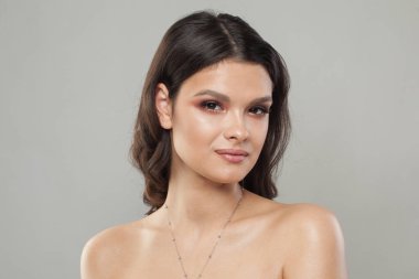 Gri stüdyo arka planında parlak parlak tenli ve koyu renk saçlı çekici genç bir kadın.