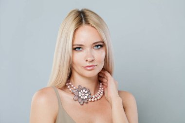 Gülümseyen manken bir kadın. Temiz, taze bir teni ve uzun sarı saçları var. Gri stüdyo arka planında takılı inci kolye takıyor.