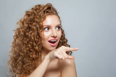 Kadın işaret ediyor. Doğal makyajlı, kıvırcık uzun saçlı ve beyaz arka planda dostça gülümseyen güzel bir kadın modeli.