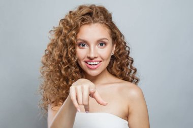 Şaşırmış bir kadın işaret ediyor. Doğal makyajlı, kıvırcık uzun saçlı ve beyaz arka planda dostça gülümseyen güzel bir kadın modeli.