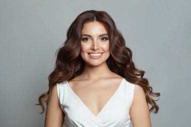 Güzel, esmer, güzel, temiz tenli, güzel, dalgalı saçlı, makyajlı ve sevimli bir kadın. Gri arka planda poz veren kameraya bakıyor.