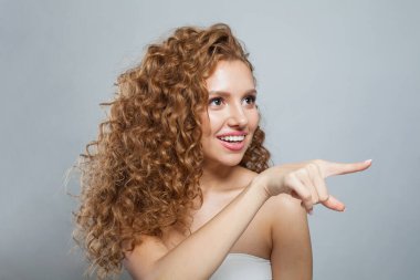 Parmakla işaret eden çekici bir kadın. Doğal makyajlı, kıvırcık uzun saçlı ve beyaz arka planda dostça gülümseyen güzel bir kadın modeli.