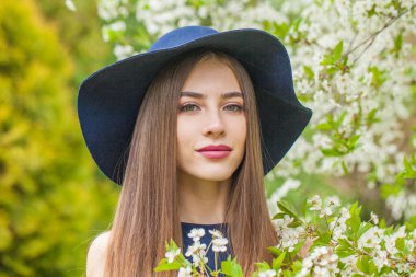 Bahar esmer kadın portresi. Uzun saçlı ve makyajlı güzel bir bayan model. Çiçek bahçesinde klasik mavi şapka takıyor.