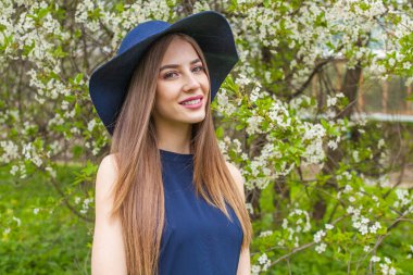 Mutlu esmer kadın portresi. Uzun saçlı ve makyajlı güzel bir kadın model. Baharın bahçesinde klasik mavi şapka takıyor.