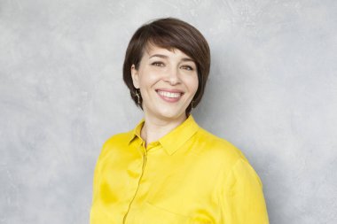 Sarı gömlekli mutlu iş kadını gri arka planda kameraya bakıyor.