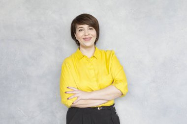 Sarı gömlekli gülümseyen iş kadını gri arka planda duran kameraya bakıyor.
