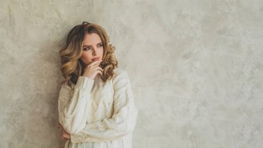 Makyajlı, dalgalı saçlı, beyaz kazaklı, gri alçı kaplama arka plan duvarlı güzel bir kadın.