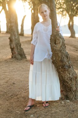 Beyaz gömlekli sarışın genç kız ormanın ve güneş ışığının arka planında ağacın yanında duruyor.