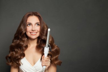 Parlak, uzun kıvırcık saçlı, kıvırcık saçlı çekici kadın saç maşasını gösteriyor. Saç bakımı, saç bakımı ve şekillendirme