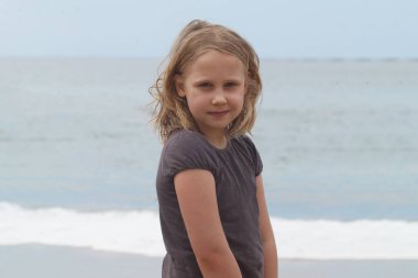 Küçük kız deniz sahilinde kameraya bakıyor.