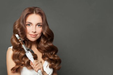 Sağlıklı esmer model, gri arka planda saç maşası kullanıyor. Kuaför, güzellik salonu ve kuaför konsepti