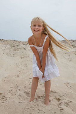 Açık havada kumların üzerinde duran beyaz elbiseli neşeli sarışın kız çocuğu..