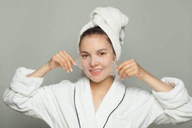 Nemlendirici, gençleştirici beyaz kumaş maskeli, beyaz arka planda kameraya gülümseyen sağlıklı genç bir kadın.