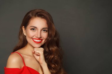 Neşeli esmer, şık makyajlı, kahverengi kıvırcık saçlı, siyah arka planda kırmızı dudaklı bir kadın. 