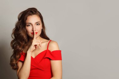 Çok çekici bir genç kadın, parmaklarını dudaklarında tutarken ve beyaz arka planda dururken sessizlik işareti yaparken sır saklıyor.